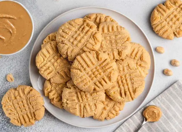 Top view of rich vegan peanut butter cookies with Himalayan Tartary Buckwheat by a bowl and spoon of peanut butter and shelled peanuts.