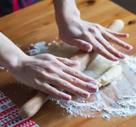 Close on a girl's hands working a rolling pin over a flattened mound of pie crust dough. 