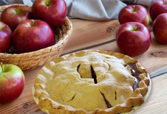 Close-up of a freshly baked Himalayan Tartary Buckwheat apple pie on a wooden table backed by Red Delicious apples.  