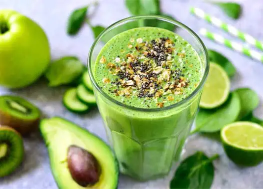 Loaded green smoothie topped with nuts and seeds and surrounded by fruits and veggies like limes, avocados, kiwi, spinach, and more.