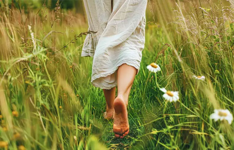 A woman walking barefoot in a grassy meadow of humus and wildflowers.