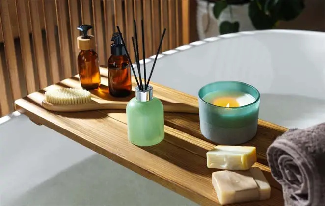 An inviting hot bath is ready to go under a stacked wooden caddy of  soaps, essential oils, a bath brush, incense sticks, and a rolled towel beside a warm soothing candle.