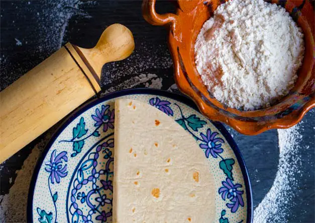 Top view of rich Himalayan Tartary Buckwheat tortillas folded on a plate by a rolling pin and rustic bowl of flour.