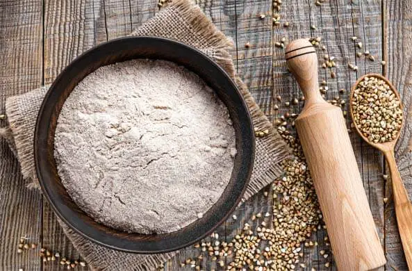 Top view of Himalayan Tartary Buckwheat flour in a bowl by a spoonful of seeds, a rolling pin, and scattered kernels on wood. 