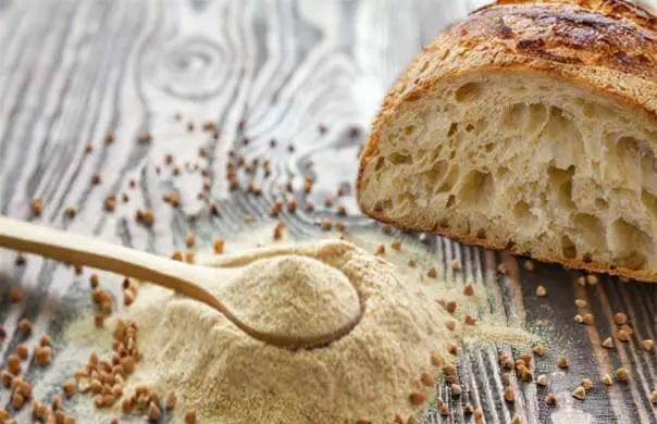 A pile and spoonful of Himalayan Tartary Buckwheat flour and scattered seeds by a sliced loaf of HTB sourdough bread on a board.