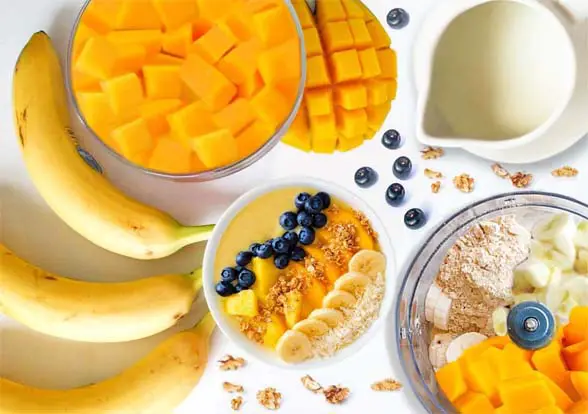 Top view of HTB Rejuvenate smoothie ingredients including mango, bananas, plant milk, a stacked blender with Himalayan Tartary Buckwheat superfood protein powder. 