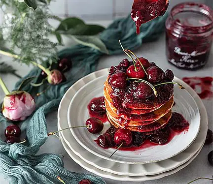 Stack of succulent Himalayan Tartary Buckwheat blinis lavished in cherries and coulis by a jar of jam and pink roses on a countertop.