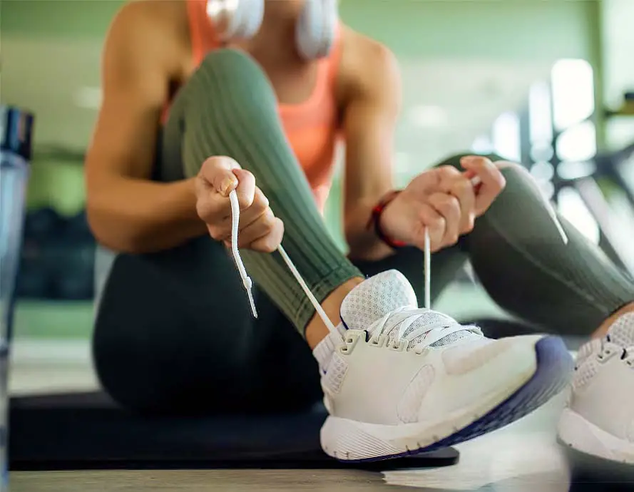 A girl in athletic clothes with headphones lacing her jogging shoes on a gym mat.