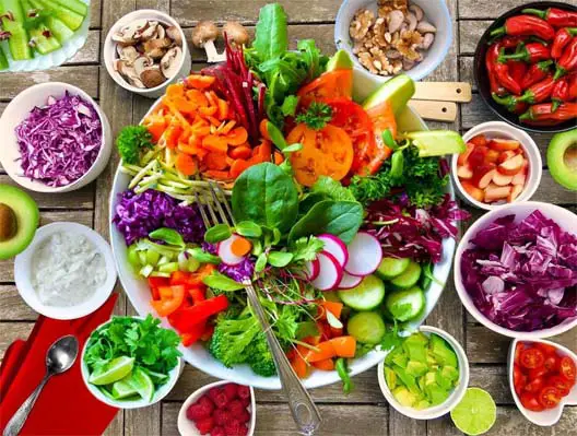 Top view of a huge bowl of mixed green salad surrounded by cups of veggies, fruit, nuts, seeds, vegan dressing, and utensils.  