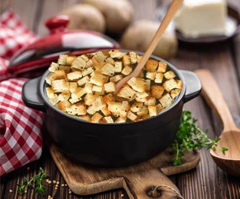 Large pot of homemade HTB sourdough croutons on a table by a checked kitchen cloth, whole potatoes, vegan butter, and fresh herbs.