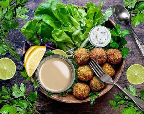 Top view of rich vegan Himalayan Tartary Buckwheat falafel balls on a bed of greens with vegan Ranch, tahini, and lemon and lime wedges. 