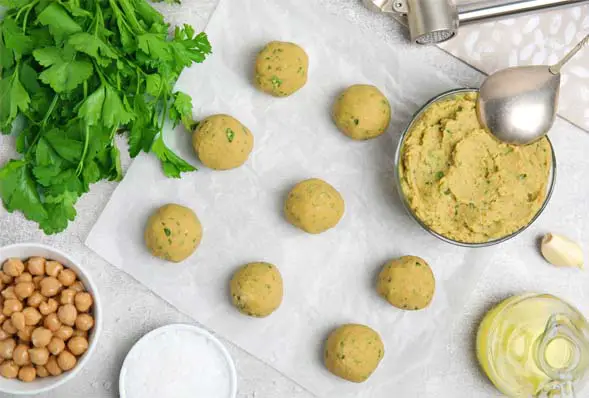 Top view of raw vegan falafel balls on parchment paper by fresh parsley, garlic, and cups of falafel mix, olive oil, chickpeas, and vegan Ranch dip.