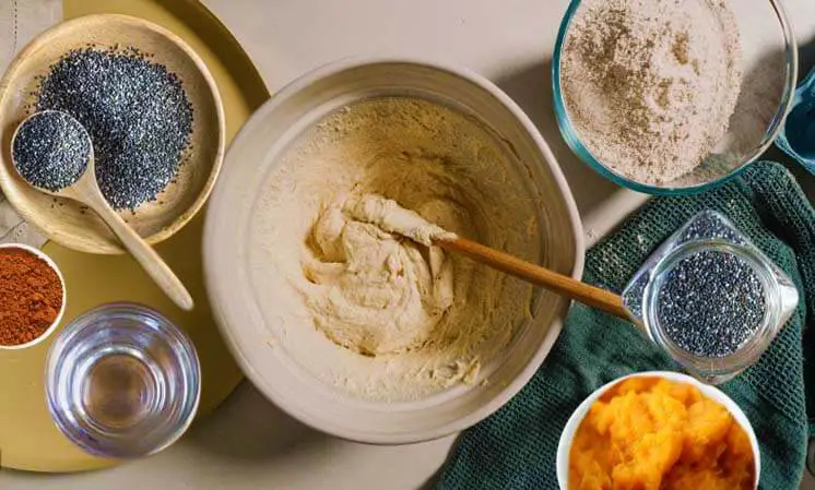 Top view of select pumpkin bread ingredients including batter in a mixing bowl, HTB flour, fresh pumpkin, chia seeds, pumpkin pie spice, and water. 