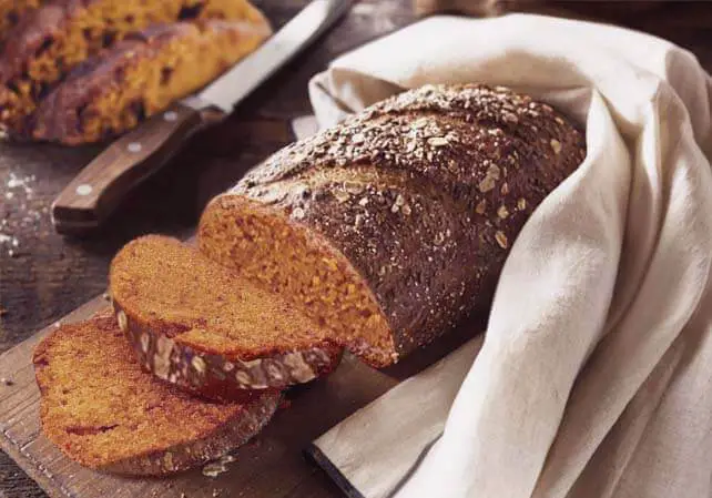 Sliced loaf of rich Himalayan Tartary Buckwheat pumpkin bread on a cutting board by a knife and hugged by a kitchen towel warmer.