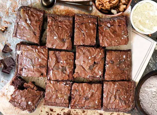 Top view of delicious vegan Himalayan Tartary Buckwheat almond flour brownies with cacao frosting on parchment paper by bowls of walnuts, almond flour, HTB flour, and chocolate chunks.