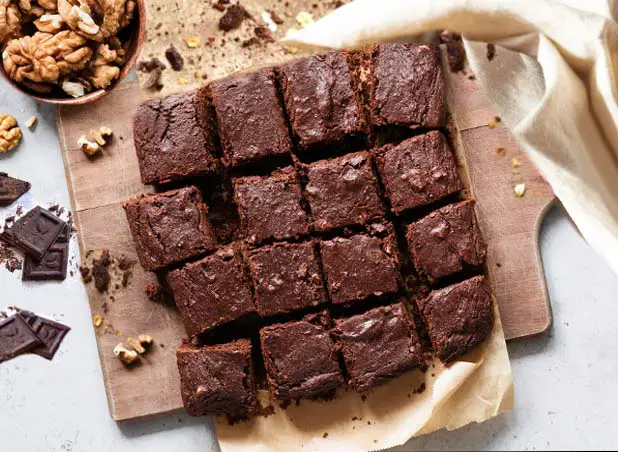 Top view of delicious vegan Himalayan Tartary Buckwheat almond flour brownies with cacao frosting on parchment paper by bowls of walnuts, almond flour, HTB flour, and chocolate chunks.