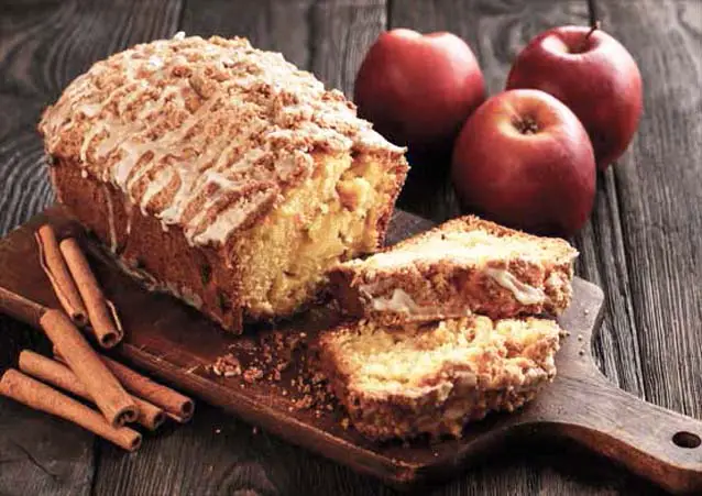 Rich and moist loaf of glazed vegan Himalayan Tartary Buckwheat apple bread sliced on a cutting board by fresh apples and cinnamon sticks.