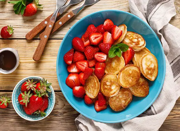 Top view of rich Himalayan Tartary Buckwheat blinis and fresh strawberries on a decorative rustic tabletop with maple syrup, utensils, and a cotton cloth.