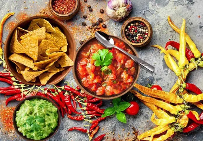 Top view of bowls of homemade salsa, chips, guacamole, and chili peppers by slews of fresh jalapenos, tomatoes, and herbs.