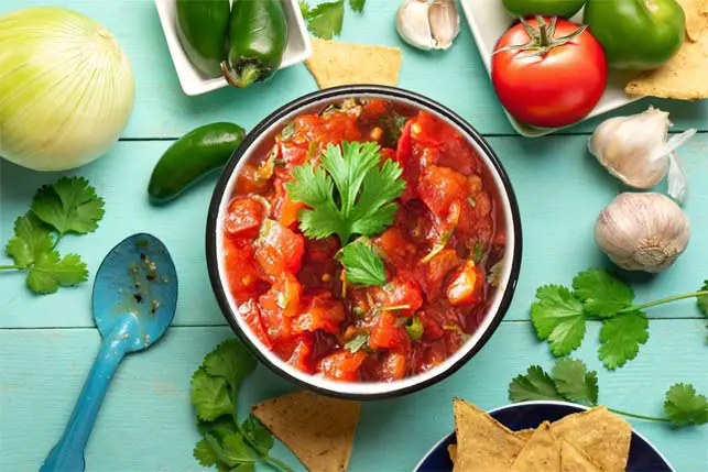 Top view of a delicious bowl of chunky homemade salsa by fresh whole tomatoes, peppers, garlic, herbs, and stone ground chips. 