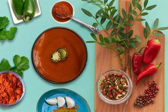 Bowl of deep red lava sauce surrounded by chilis, garlic, spices, and greens.  