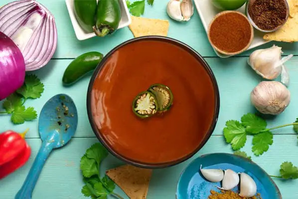 Top view of rich vegan lava sauce surrounded by peppers, garlic, onions, and herbs.