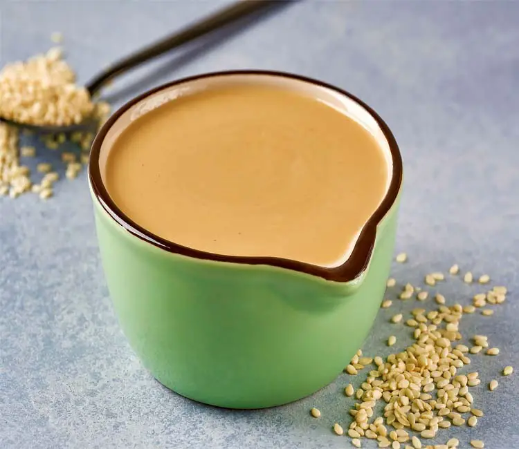 Close-up of a rich creamy mug of homemade vegan tahini by a spoonful of sesame seeds. 