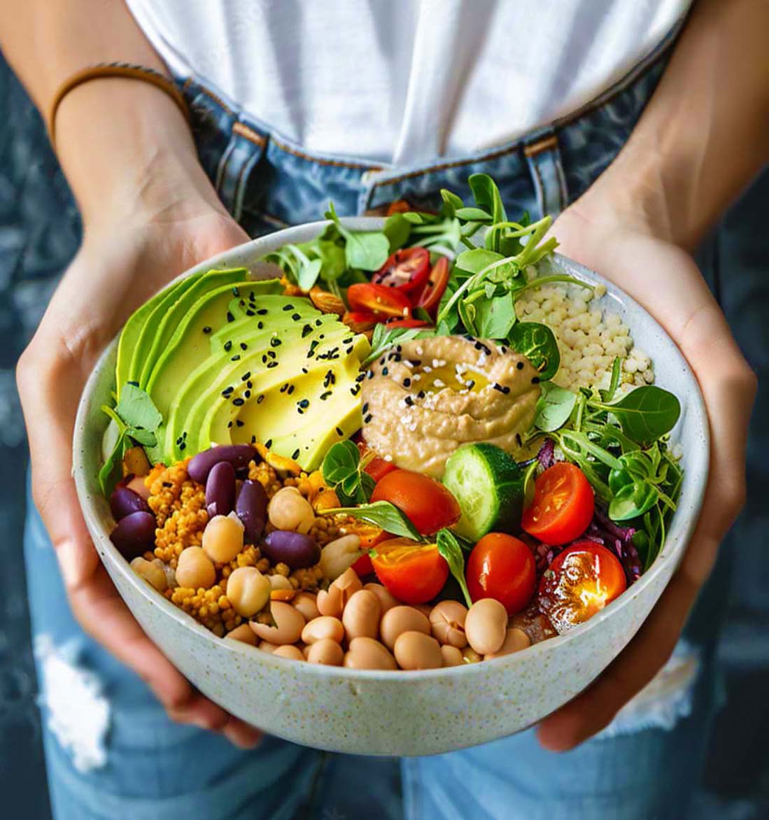 A woman's hands holding a loaded organic buddha bowl packed with fruits, veggies, grains, legumes, and fresh tahini dip. 