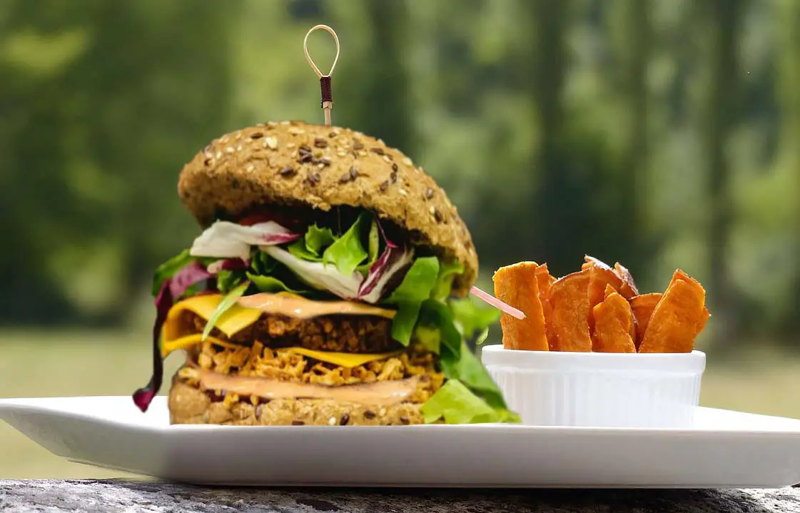 Stacked vegan volcano burger with veggies, lava sauce, and a side of sweet potato fries on a picnic table. 