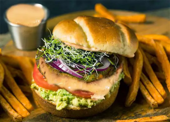 Stacked vegan Volcano Burger packed with veggies and sauce by crispy fries on a rustic board.