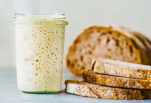 A bubbly jar of wild yeast sourdough starter by slices of crusty homemade sourdough bread.