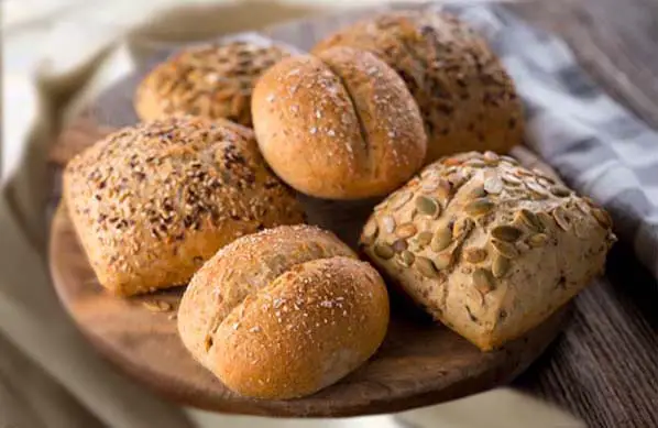 A stacked wooden platter of homemade sourdough dinner rolls with assorted nuts and seeds.