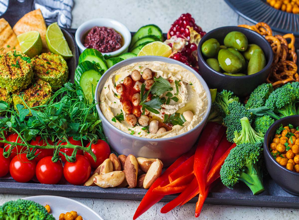 Rich bowl of homemade vegan hummus on an appetizer platter with crudite, falafel, flatbread, pomegranate, pretzels, and herbs.