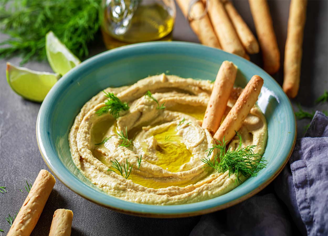 Top view of delicious homemade hummus swirled in a bowl with herbs, lime, olive oil, and organic breadsticks.