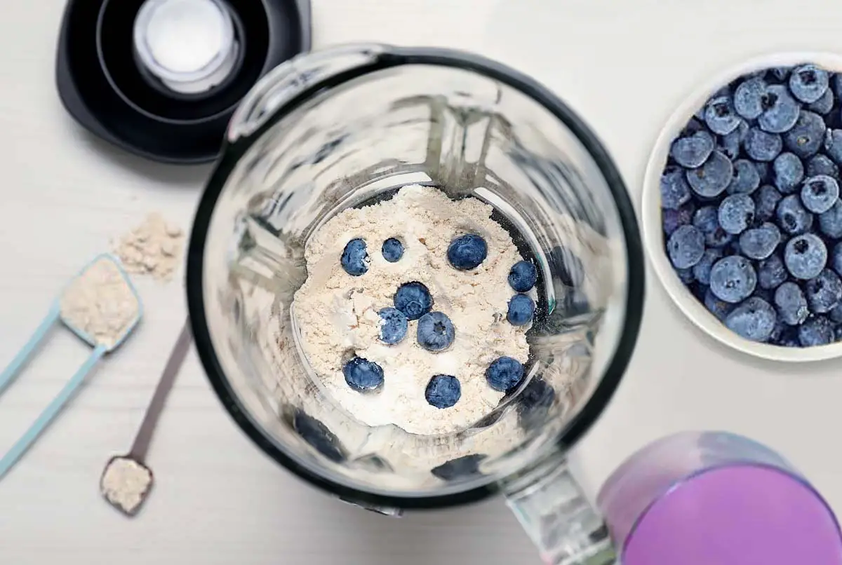 Top view of blender stacked with protein powder and blueberries by spoons of superfood, a smoothie, and blueberries in a bowl.