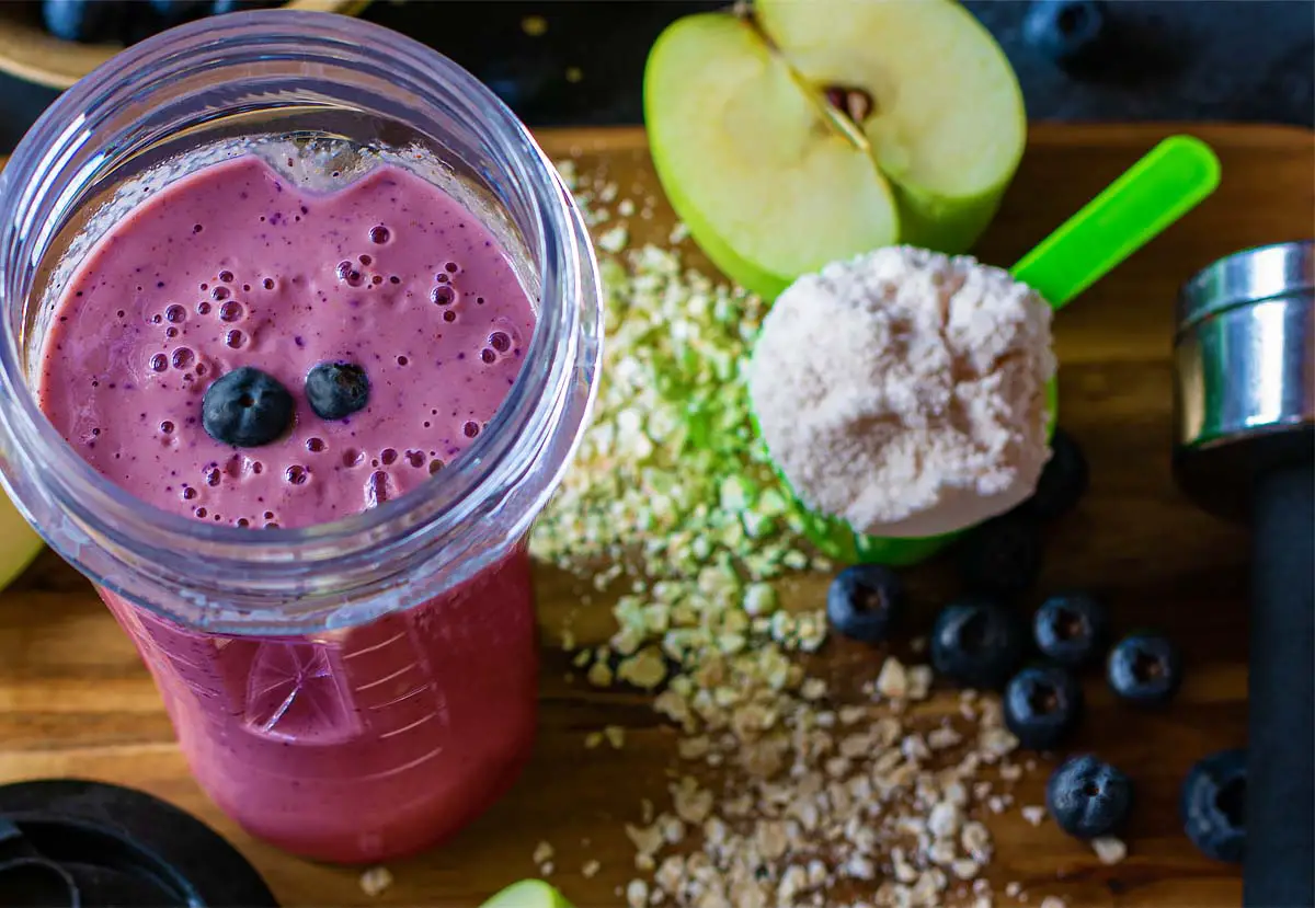 Close-up of rich vegan blueberry smoothie by a scoop of protein powder, hand weights, green apple, and organic steel cut oats. 