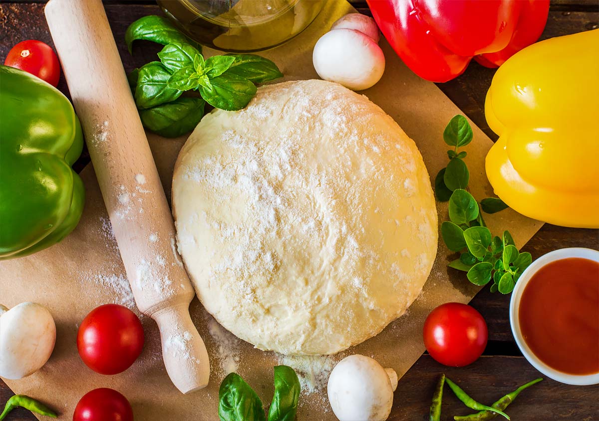 Top view of Himalayan Tartary Buckwheat pizza dough in a ball surrounded by tomatoes, garlic, mushrooms, bell peppers, greens, pizza sauce, and a rolling pin.