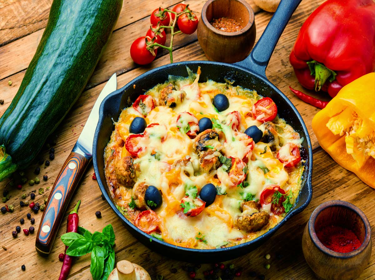 Top view of deep dish vegan pizza in a cast iron pan by fresh bell peppers, herbs, zucchini, jalapenos, peppercorns, and rich cherry tomatoes on the vine.   