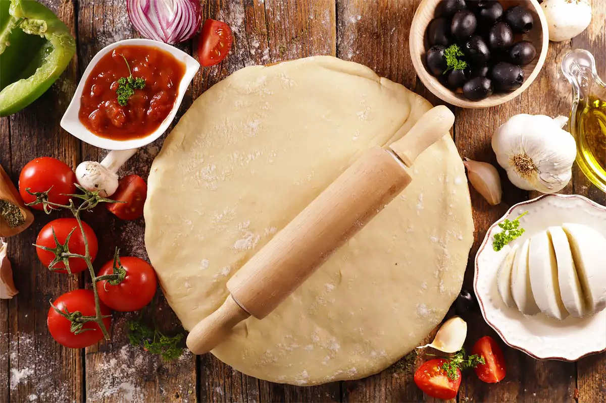 Top view of flattened pizza dough beneath a rolling pin on a board by olives, tomato sauce, garlic, bell pepper, red onion, olive oil, and more.   