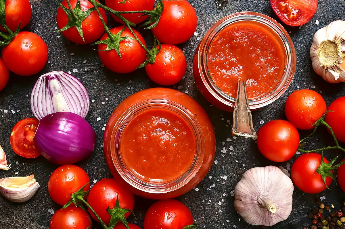 Top view of open jars of rich pizza sauce by fresh tomatoes on the vine, garlic, onions, and herbs. 