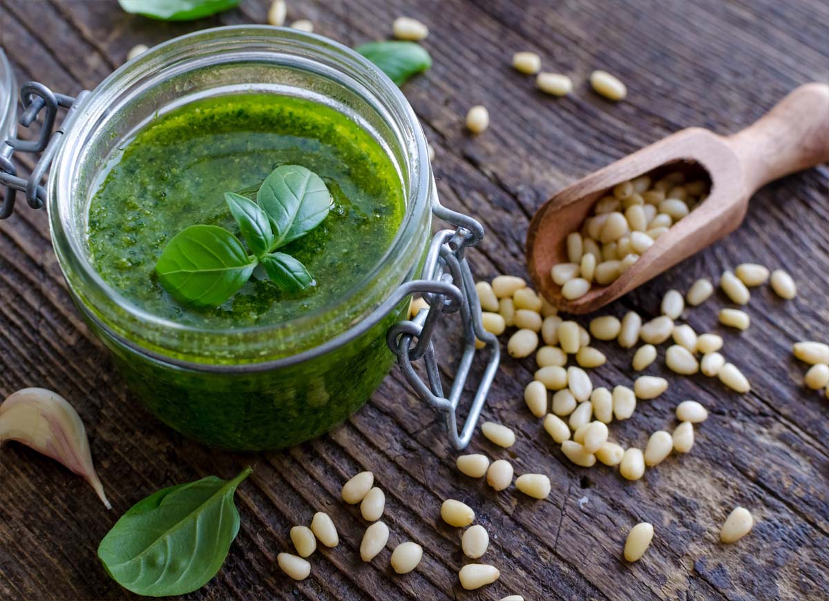 Open mason jar of rich vegan pesto by an overflowing scoop of pine nuts on a rustic board.  