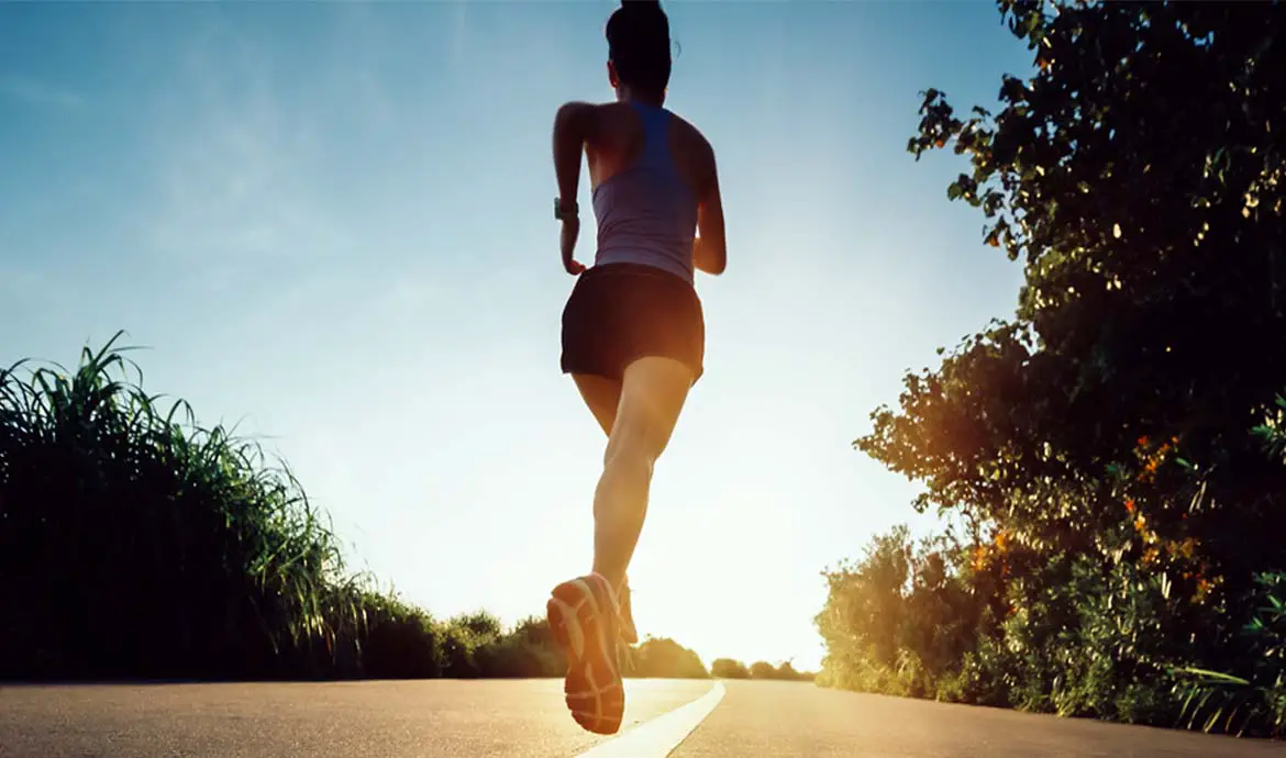 Rear view of a female runner in shorts and a tank top sprinting up a hill on a sunny country road.