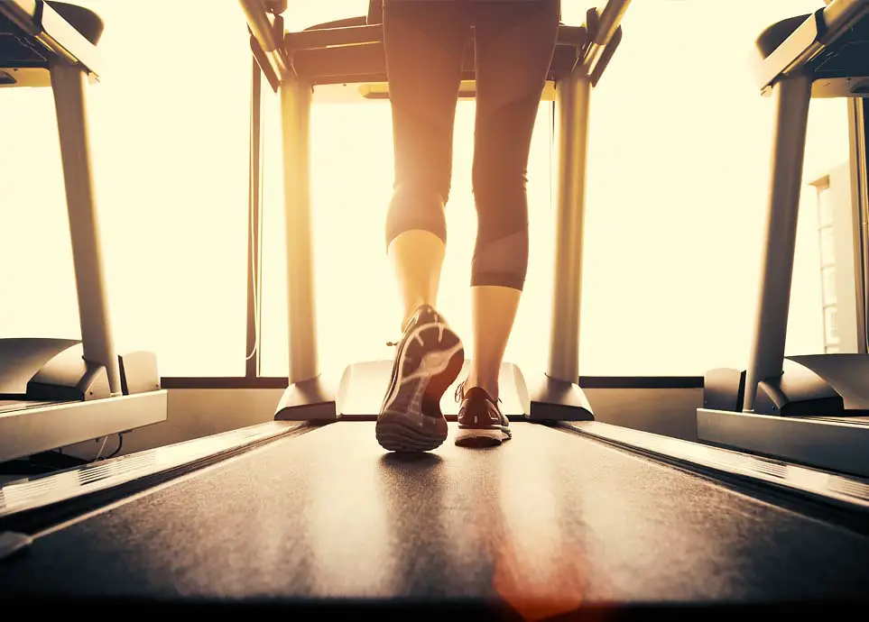 Rear view of a girl's calves walking on a gym treadmill by a sunny window.