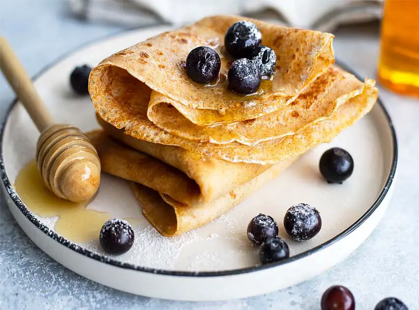 Stack of folded vegan buckwheat crepes topped with fresh blueberries and maple syrup.