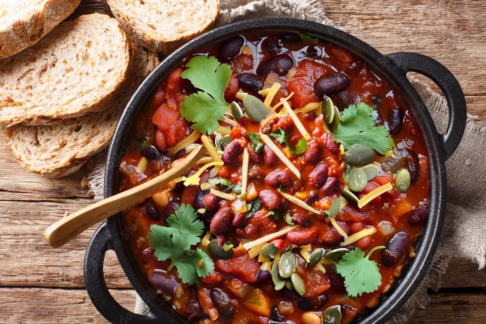 Top view of sprouted three-bean vegan chili in a pot with herbs, pumpkin seeds, and vegan cheese shreds by some crusty homemade bread slices.  