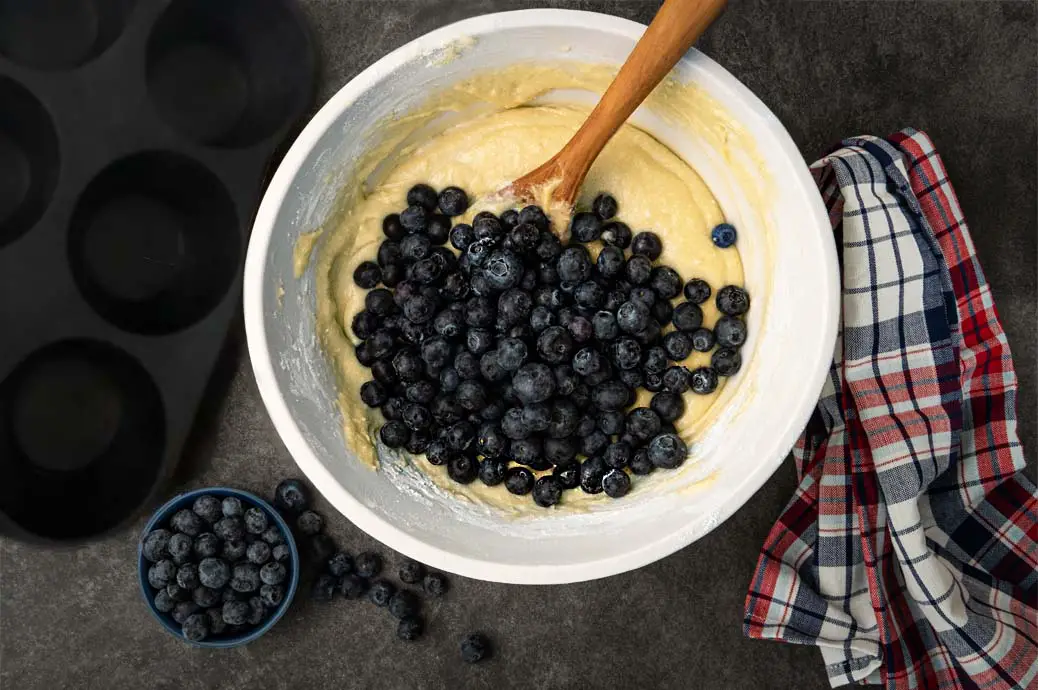 Top view of vegan blueberry muffin batter stacked with fresh berries by a cast iron muffin pan. 