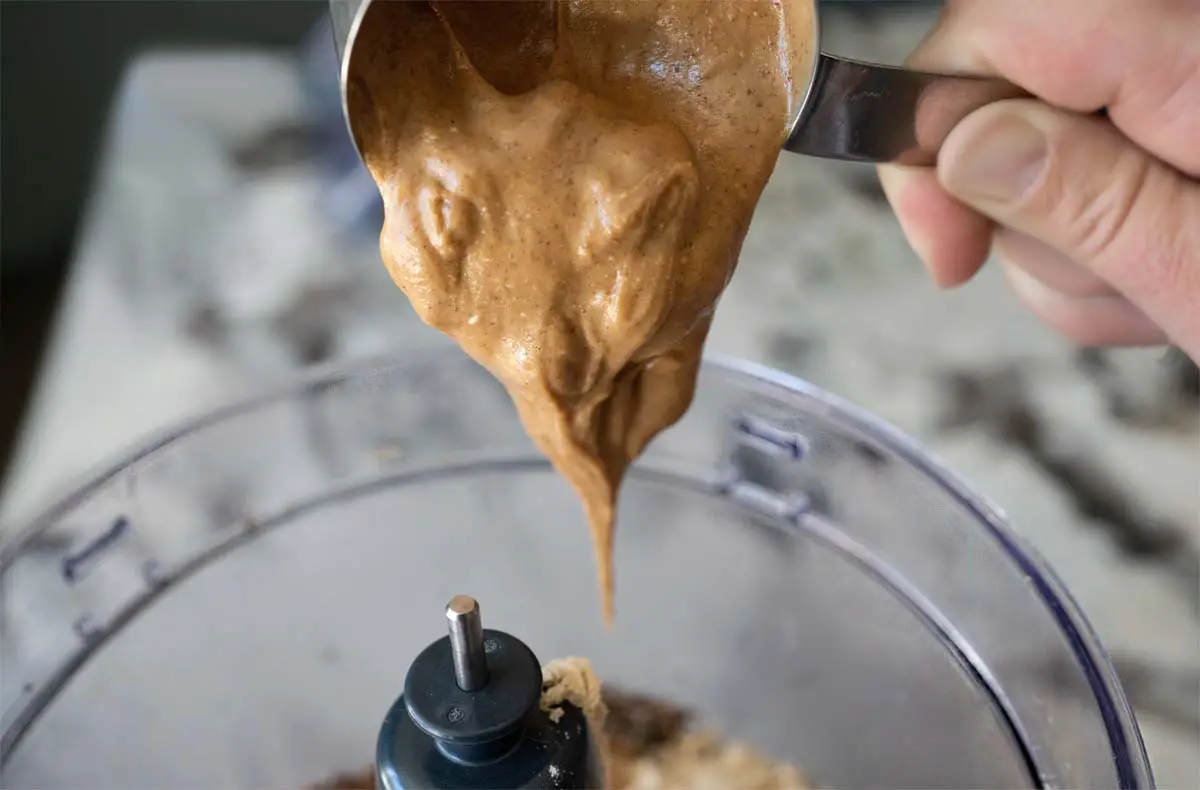 A man's hand tilts a scoop of peanut butter above a blender packed with peanut butter banana smoothie ingredients.