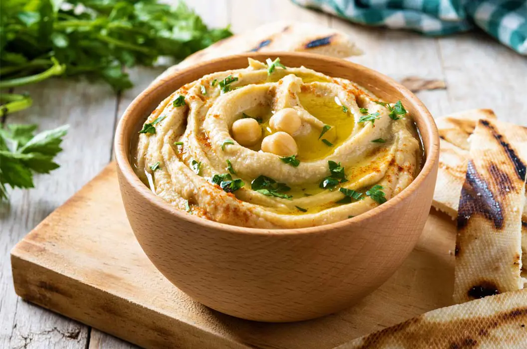 Rich and succulent bowl of organic vegan hummus and pita on a cutting board topped with chickpeas, paprika, green onions, and extra-virgin olive oil. 