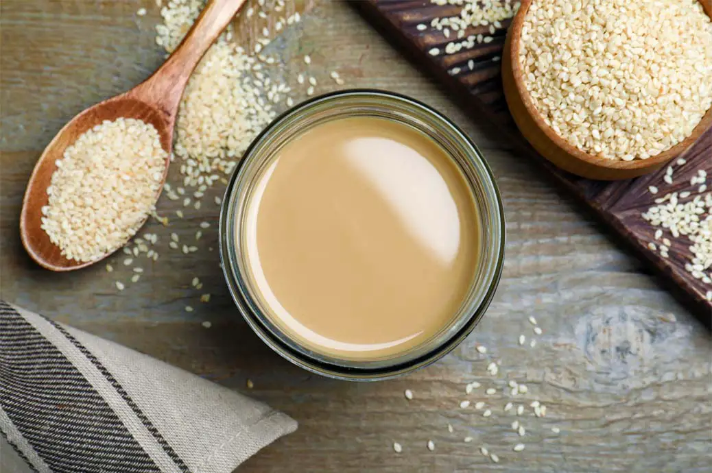 Top view of creamy rich tahini sauce by an overflowing bowl and spoonful of organic sesame seeds. 