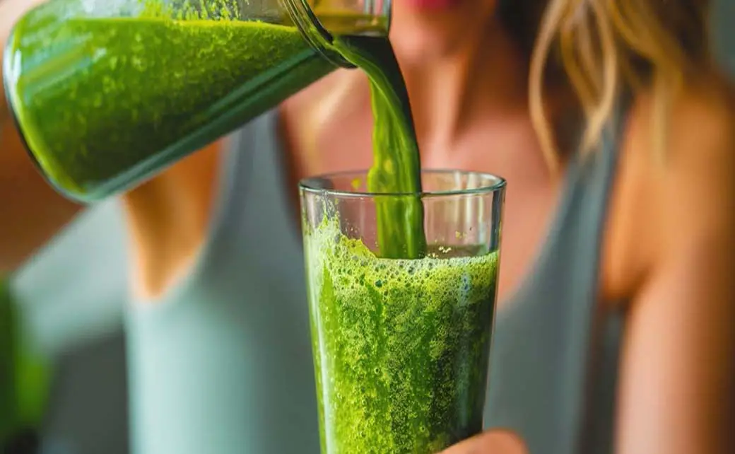 A young woman pours a green smoothie from a pitcher into a tall glass.